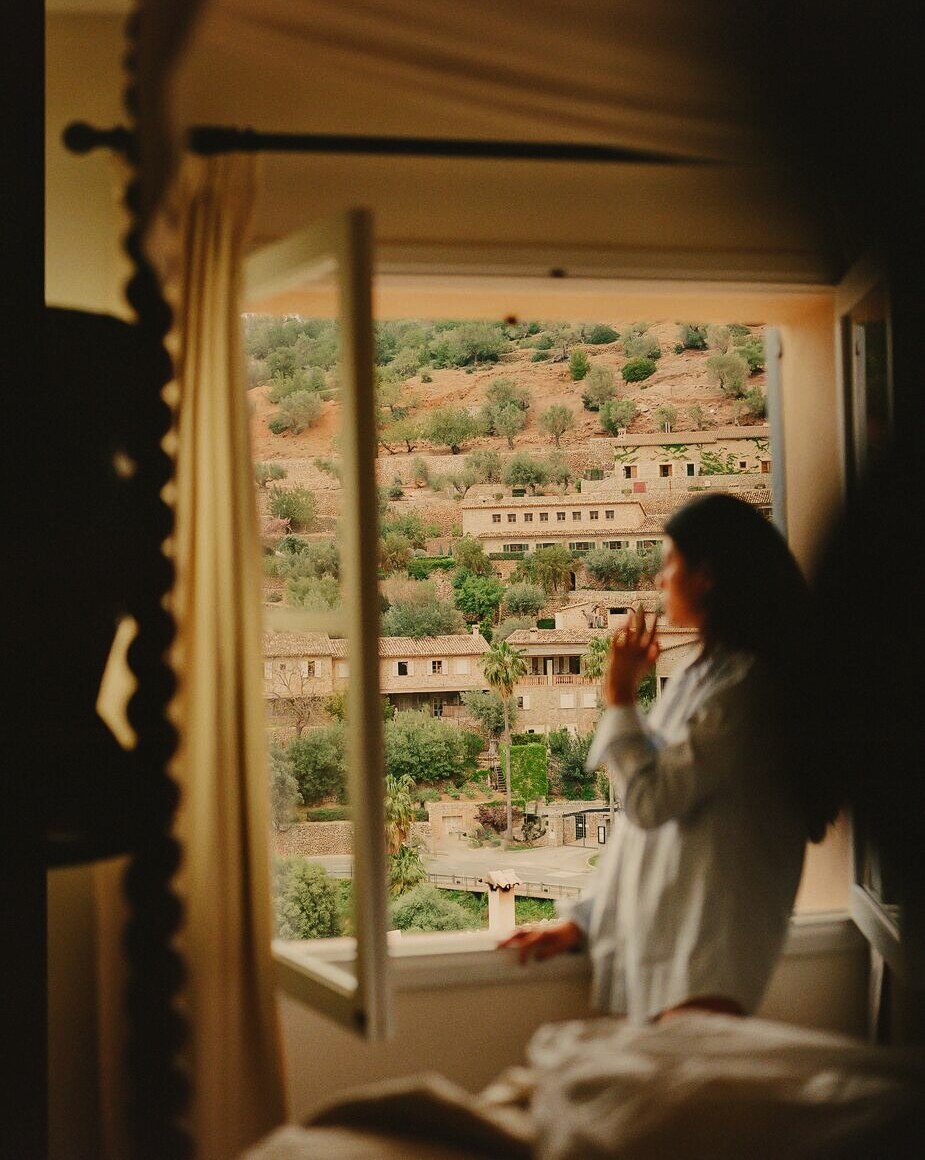 A guest in a white shirt at an open window, looking out over a hillside village