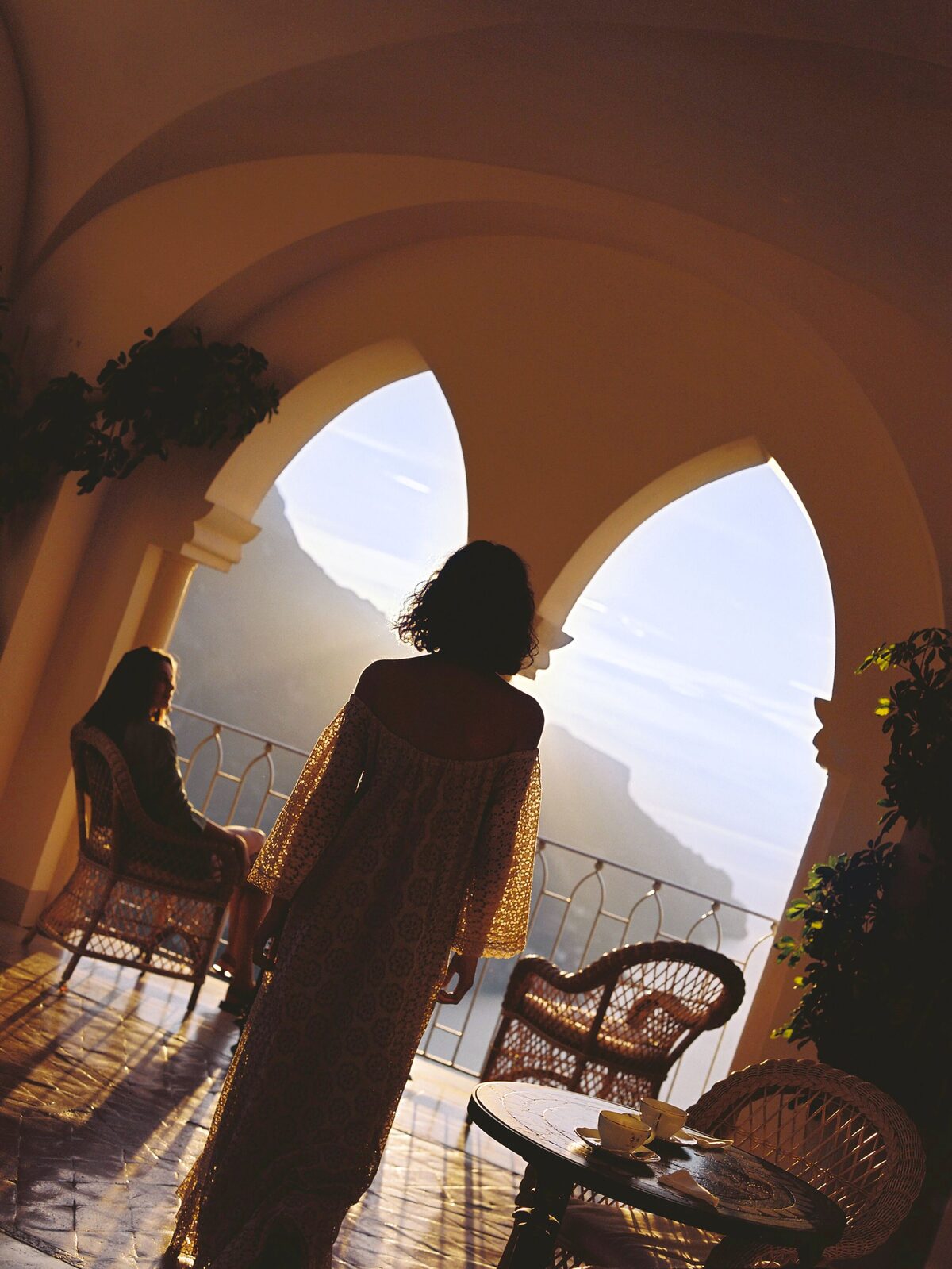Two guests on a sunlit terrace between arches, the coast beyond