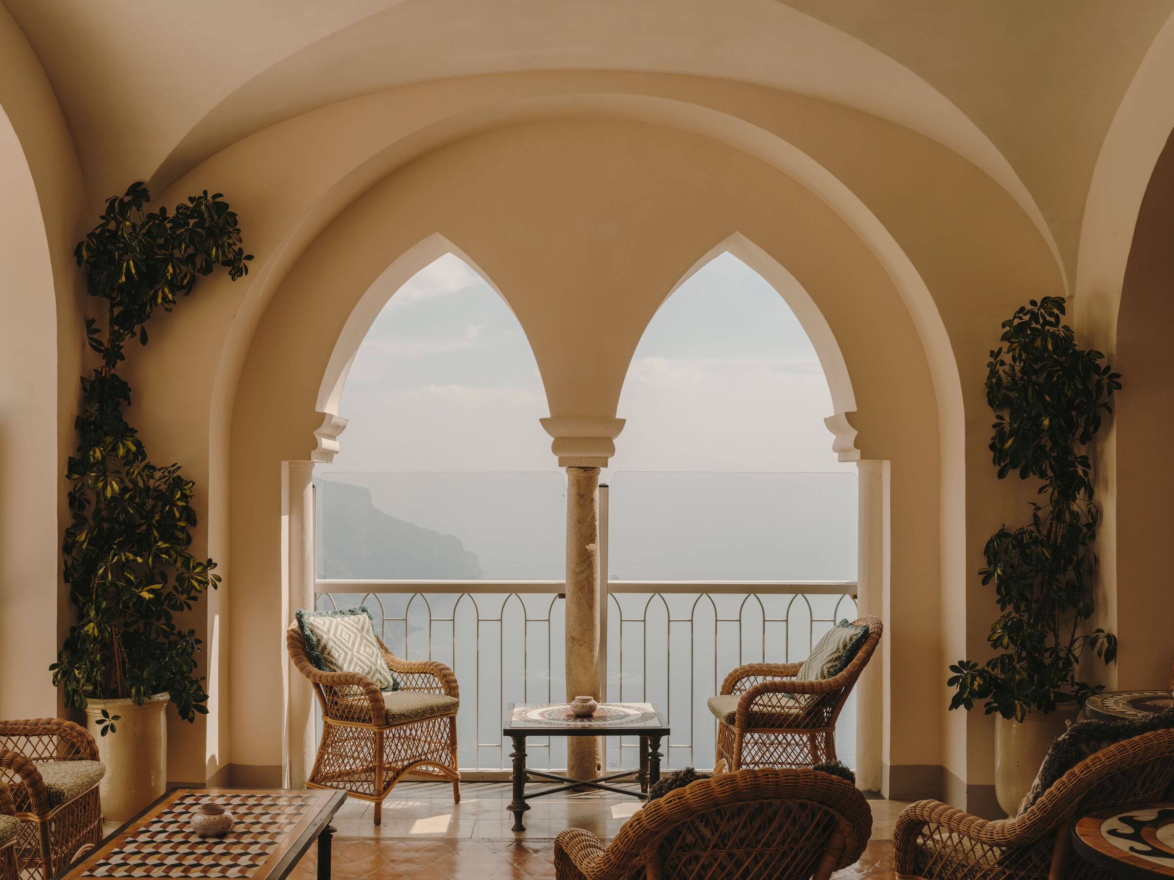A vaulted loggia with twin arches open to the sea, wicker chairs around a low table