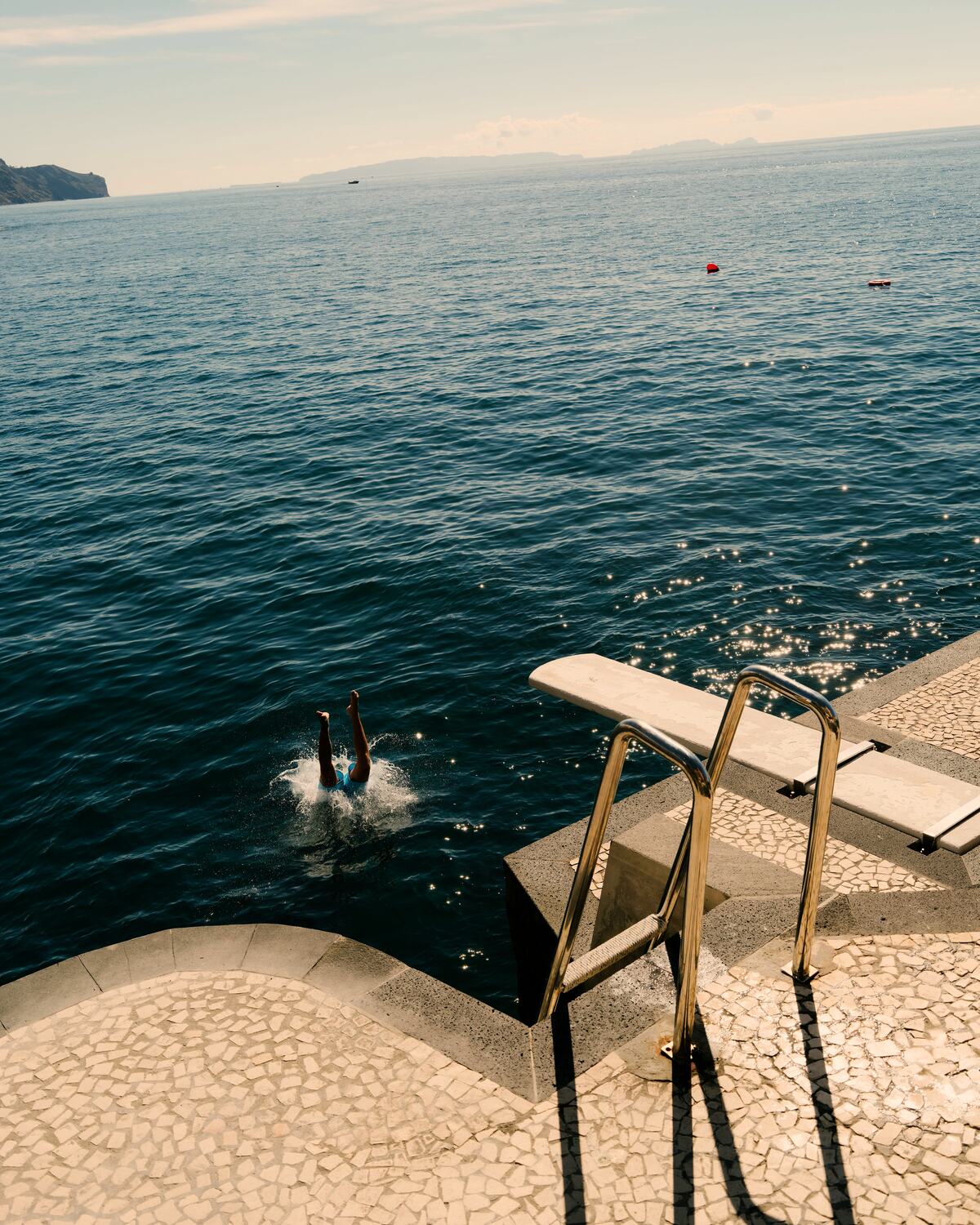 A swimmer diving into a deep blue sea from a stone jetty, only their legs above the water
