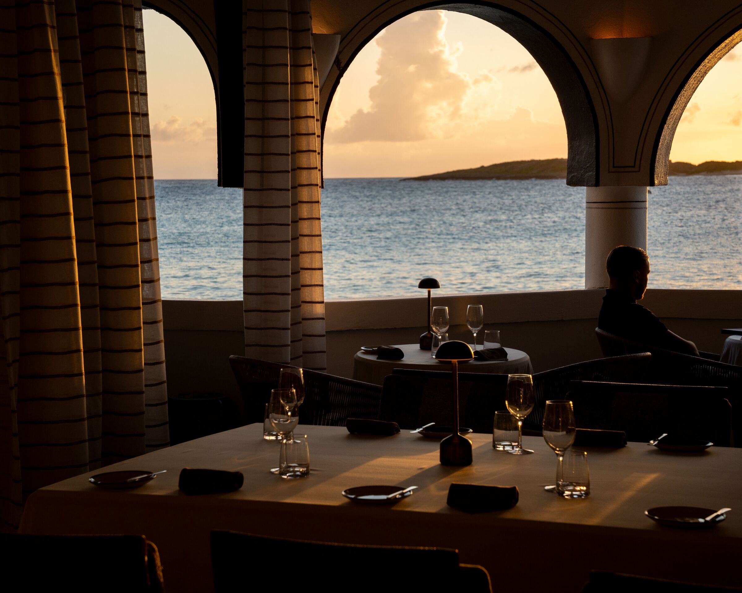A restaurant at sunset, arches open to the sea and an island on the horizon, a lone diner at the window