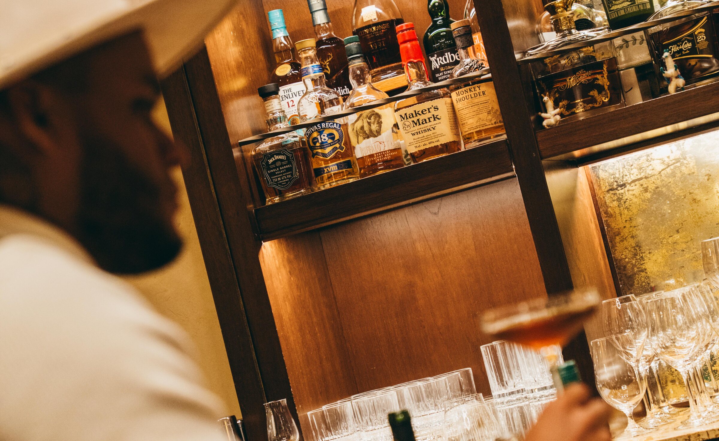 A guest in a straw hat at the bar, bottles lit behind