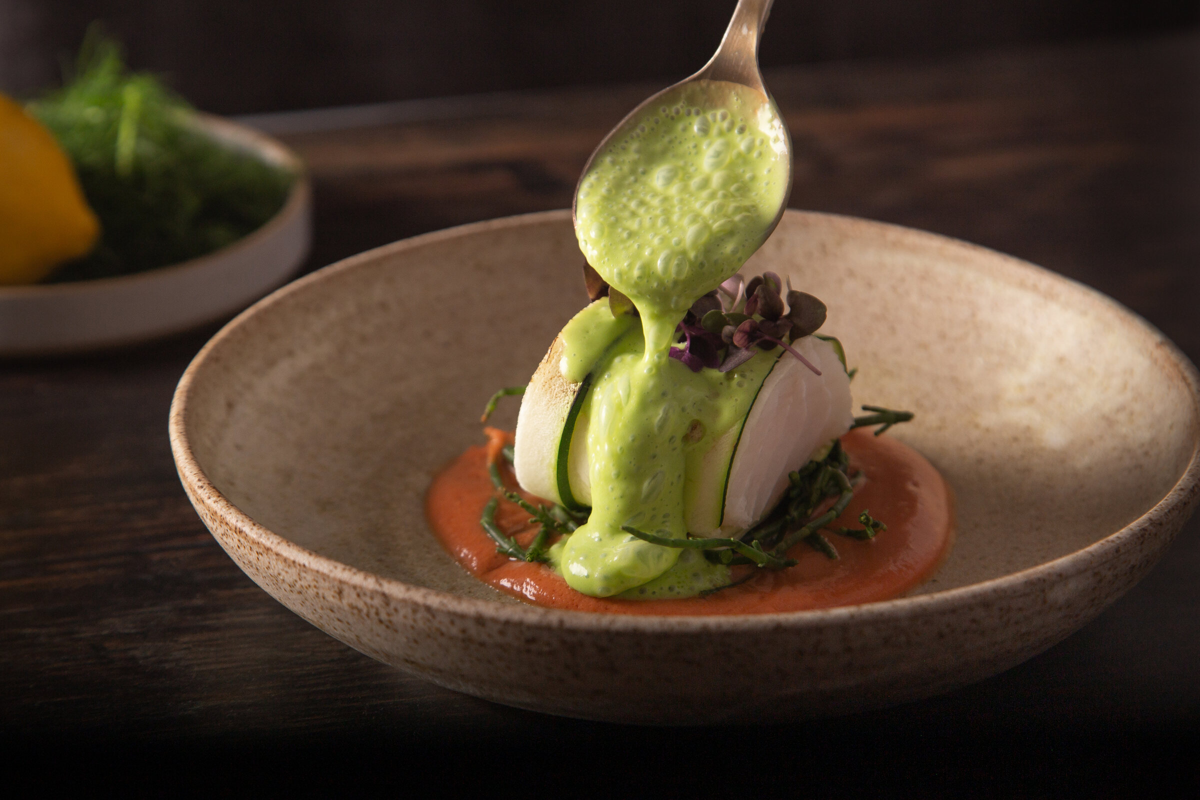 A spoon of green herb sauce poured over a courgette-wrapped dish in a stone bowl