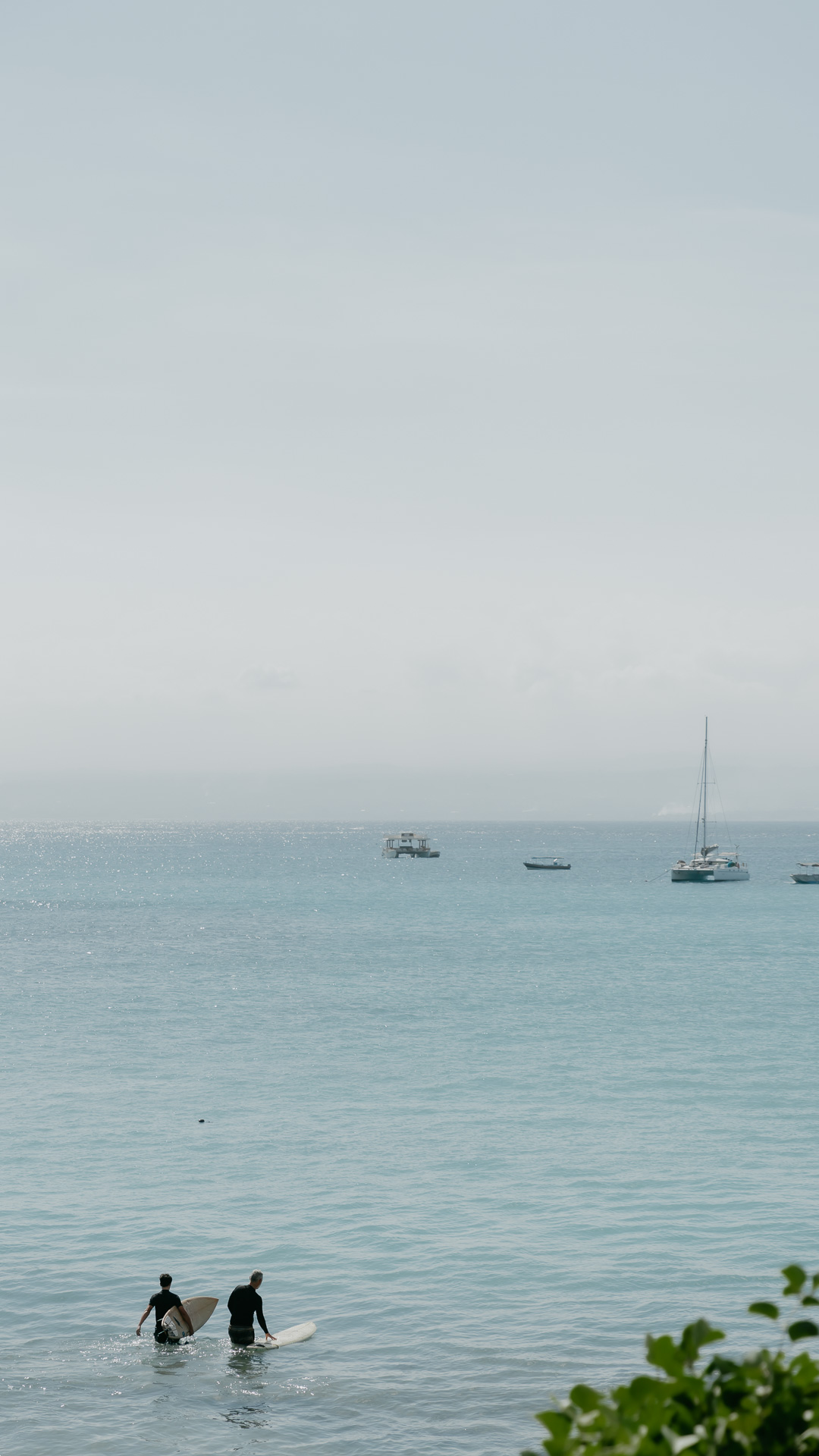 Two surfers wading into the calm bay below Zambala, boats moored beyond
